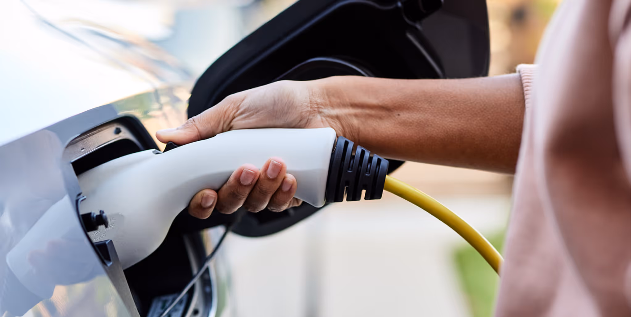 Person plugging a white electric vehicle charging connector with a yellow cable into a car.