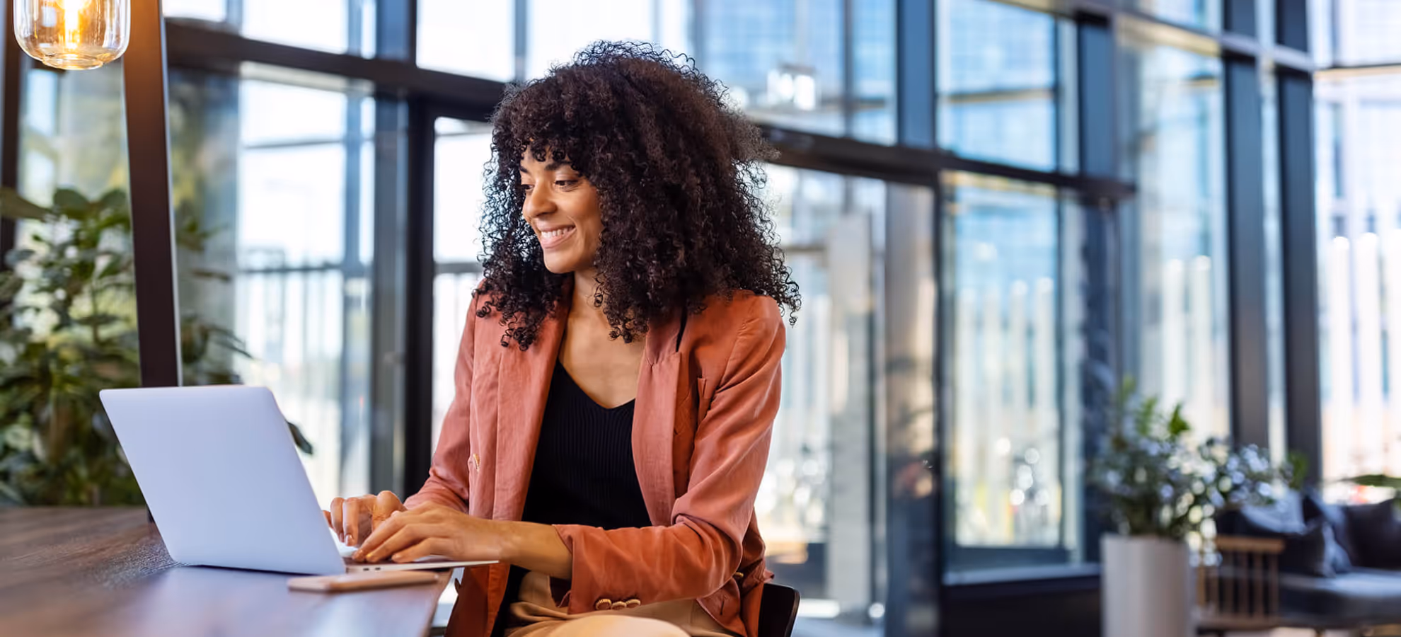 Smiling woman with curly hair working on a laptop at a wooden table in a modern office with large windows.