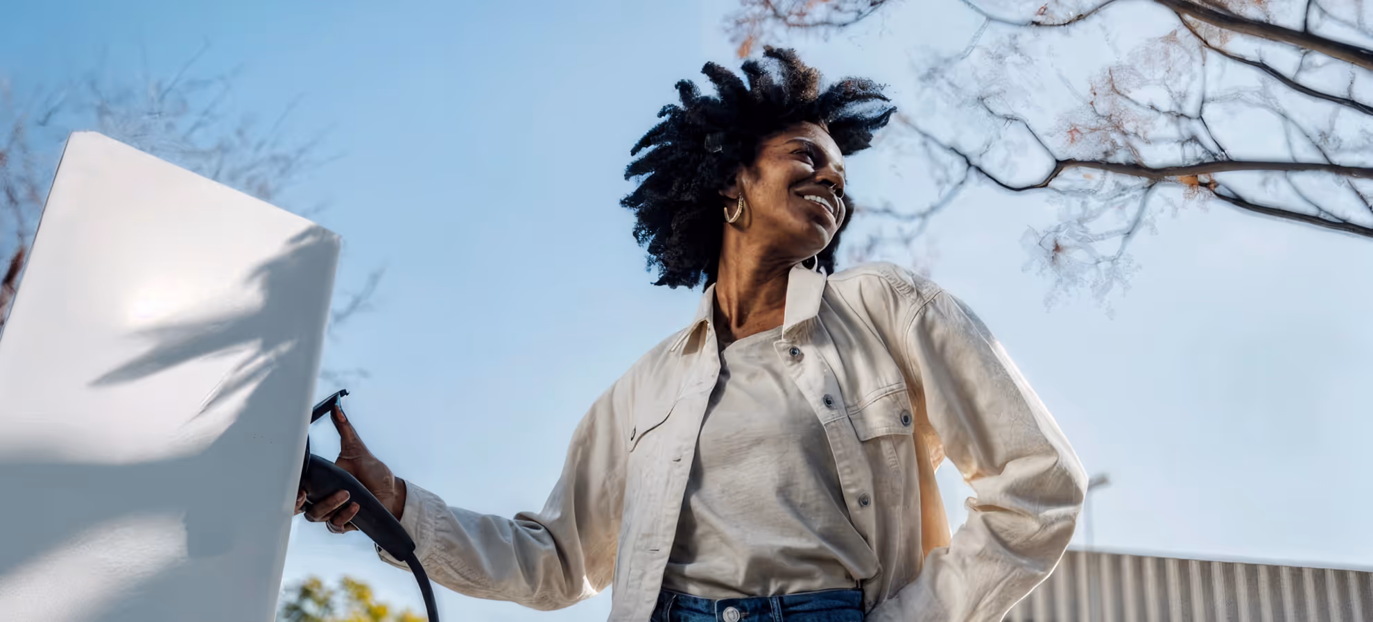 Smiling woman with natural hair holding a charging plug near an electric vehicle outdoors.
